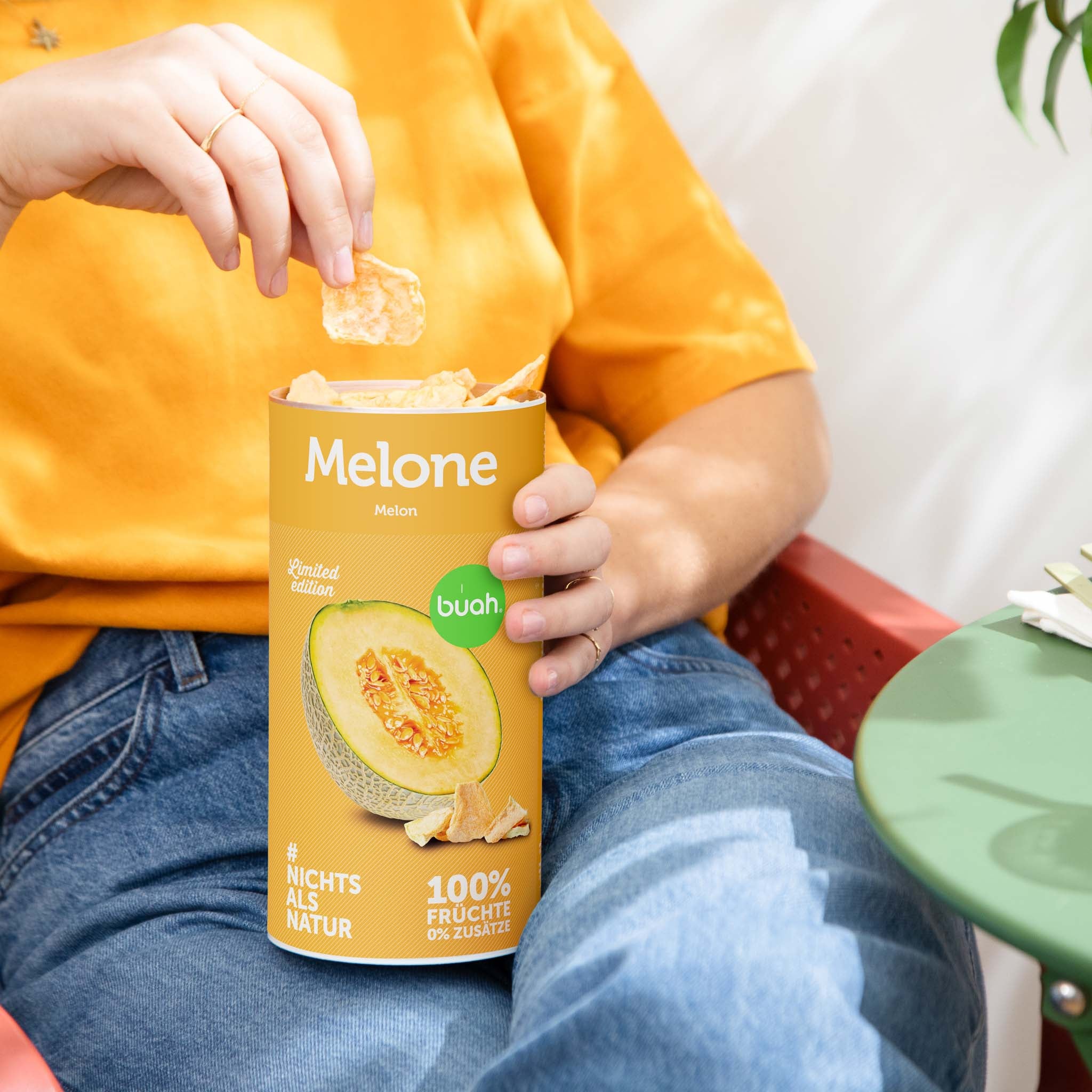 buah Dose gefriergetrocknete Cantaloup Melone steht auf dem Schoss einer jungen Frau in orangenem Shirt, die in die Dose greift.