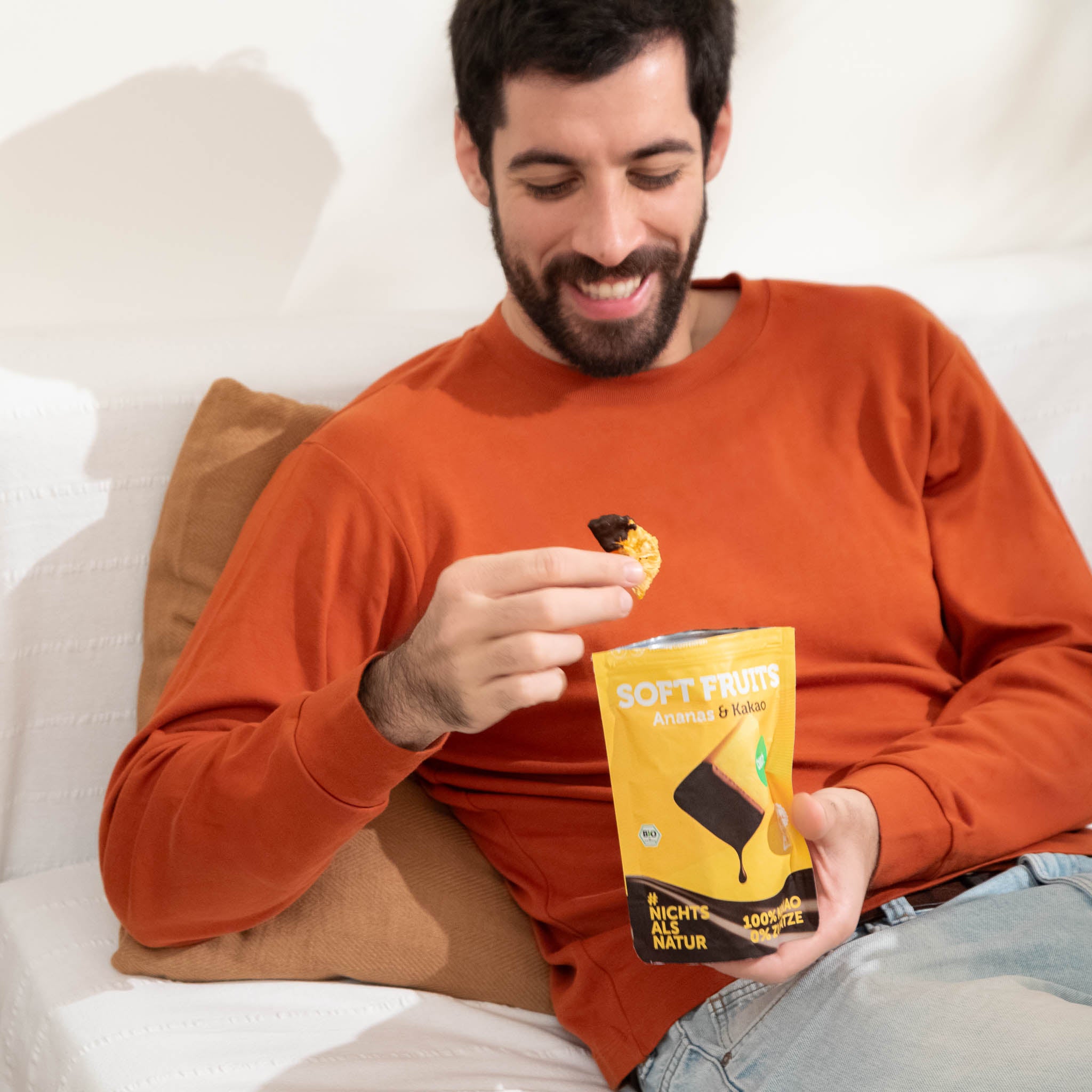 Ein lĂ€chelnder Mann im orangefarbenen Pullover sitzt gemĂŒtlich auf einem weiĂen Sofa und isst einen schokolierten Fruchtsnack. In der anderen Hand hĂ€lt er einen geöffneten gelben Beutel buah Soft Fruits der Sorte Ananas & Kakao.