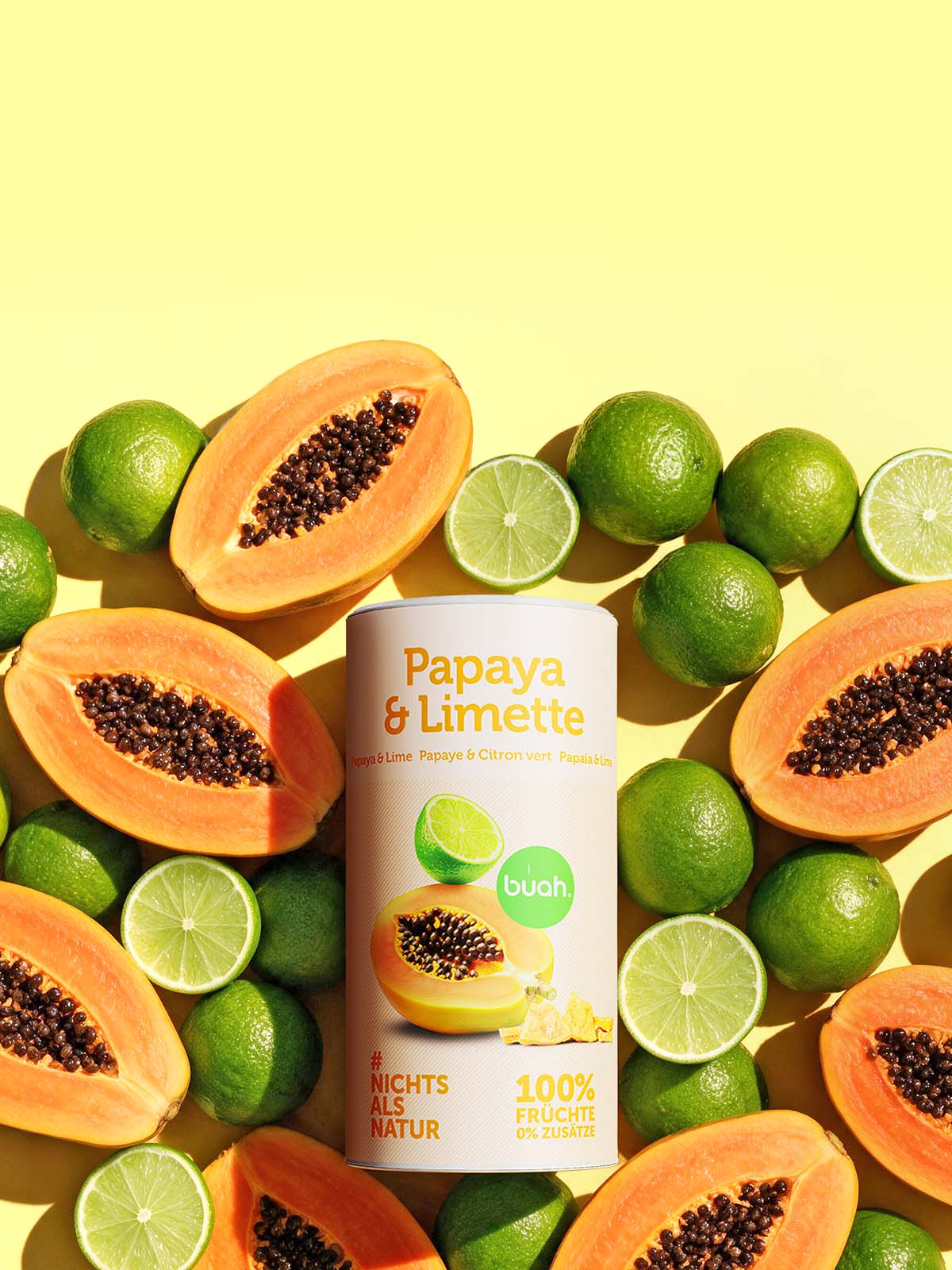 Hochformatige buah-Dose „Papaya & Limette“ mit gefriergetrockneter Papaya und Limette, umgeben von Papayahälften und Limetten auf gelbem Hintergrund.