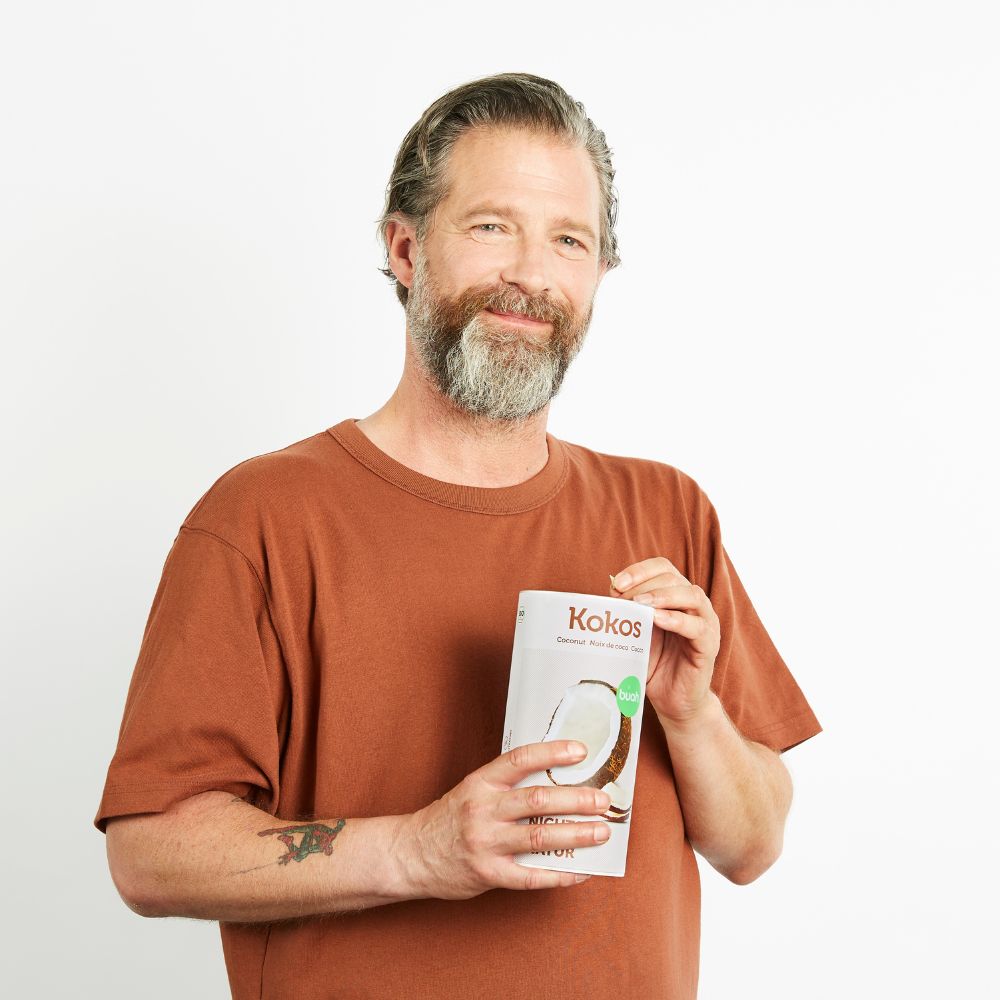 Mann Mitte 40, best Ager,  mit braunem Shirt und grau meliertem Bart isst Kokoschips aus einer Dose buah gefriergetrockneter Kokosnuss