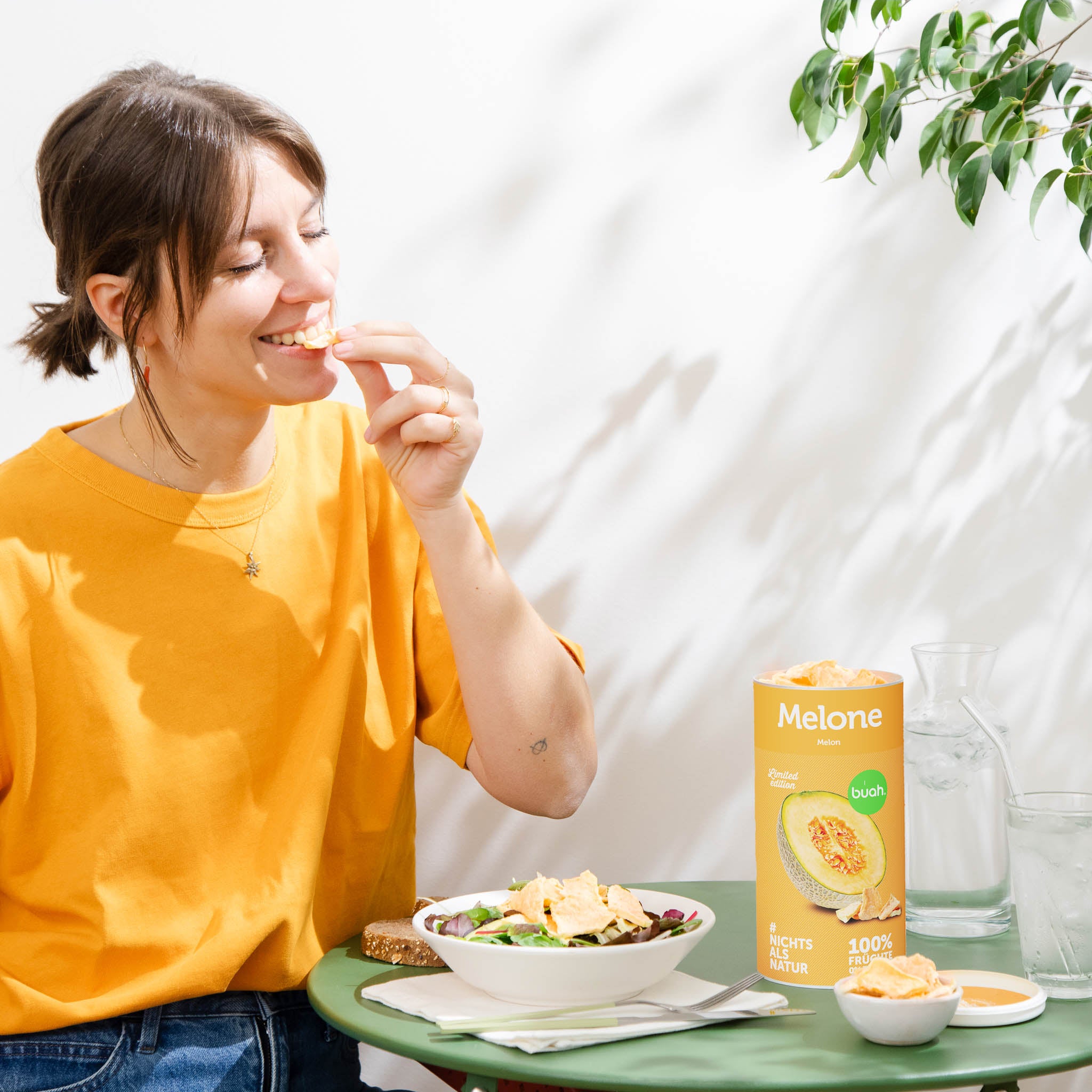 Junge Frau mit orangenem Shirt sitzt an kleinem Tisch und geniesst buah gefriergetrocknete Melone aus der Dose und gibt Melone auf Ihren Salat. 
