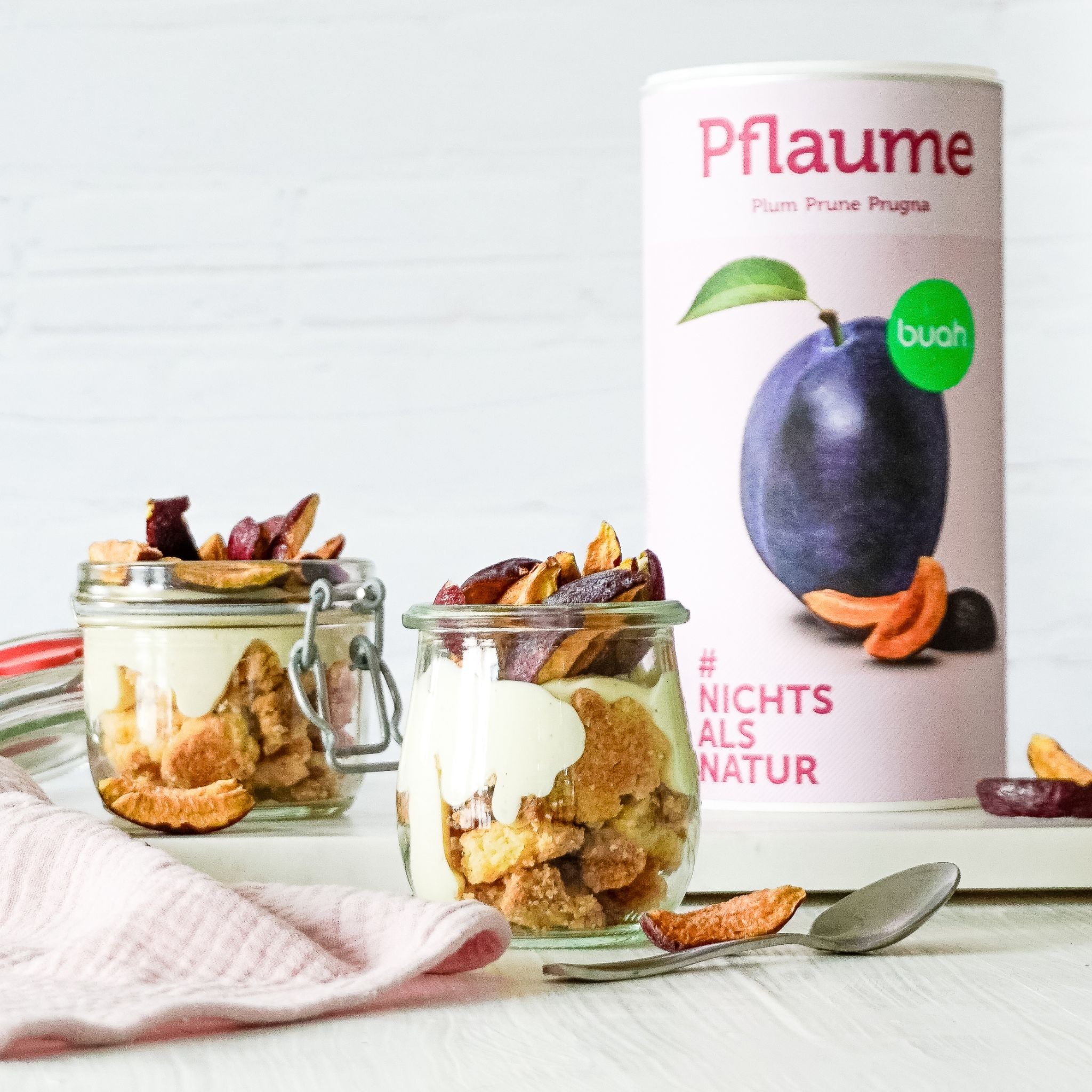 buah Dose gefriergetrocknete Pflaume im Hintergrund, davor zwei kleine Einmachgläser mit Crumble, buah Pflaume und Creme