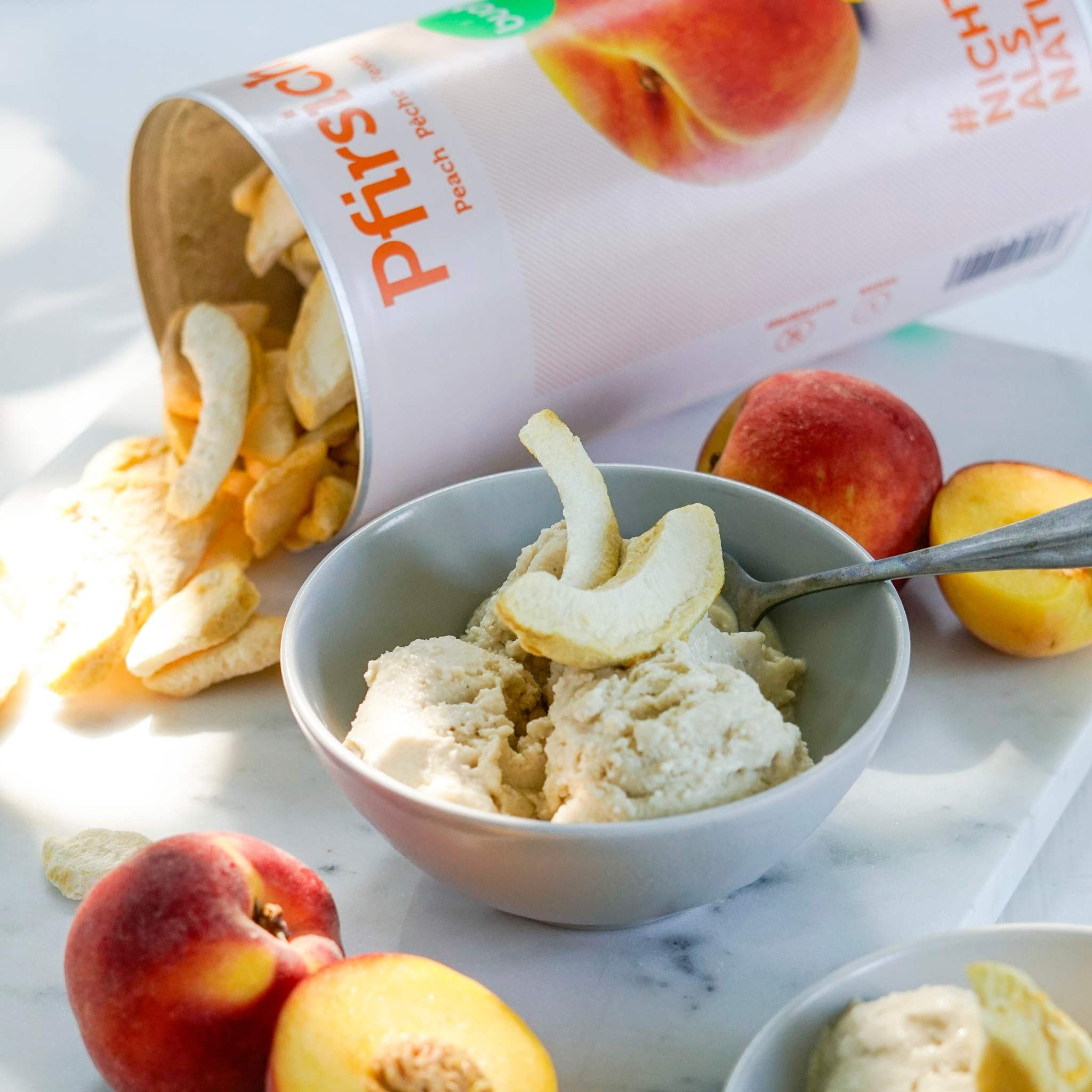 Eine graue Schale mit Eiscreme ist mit knusprigen Pfirsichstücken garniert. Dahinter liegt eine umgekippte große Dose buah gefriergetrocknete Früchte der Sorte Pfirsich, aus der weitere Fruchtstücke auf eine helle Marmorplatte rieseln. Zur Dekoration liegen frische Pfirsiche daneben.