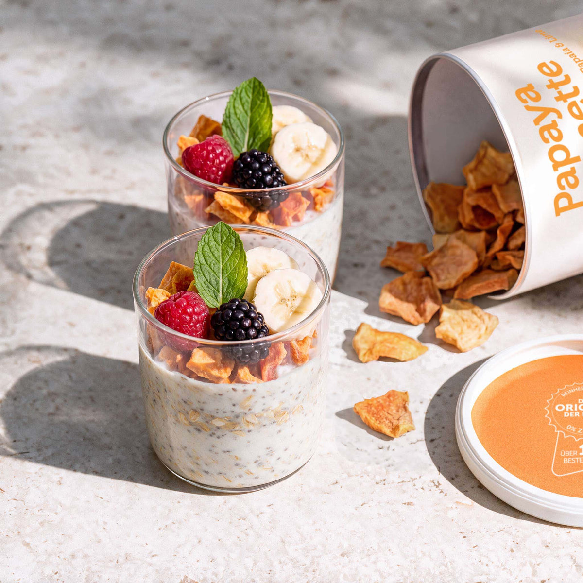 Zwei Gläser mit Joghurt und Haferflocken, belegt mit gefriergetrockneter Papaya, Beeren, Banane und Minze; daneben eine geöffnete buah-Dose Papaya & Limette.
