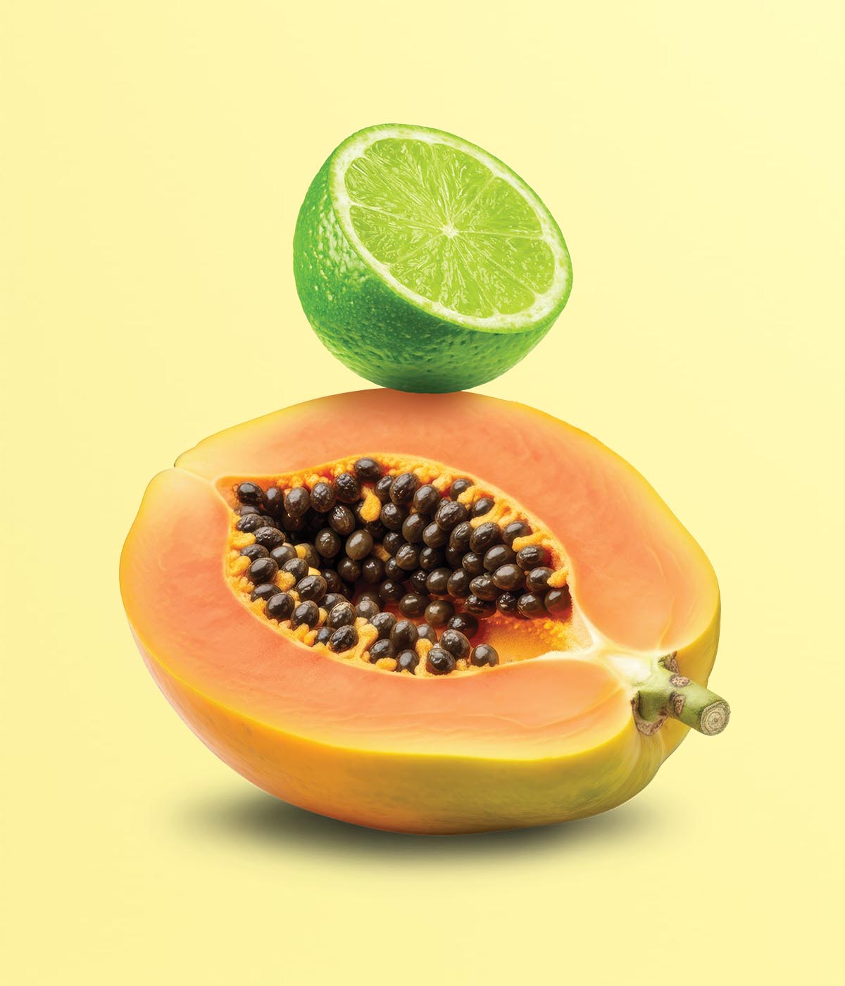 Halbe Papaya mit schwarzen Kernen und eine halbierte Limette auf hellgelbem Hintergrund.